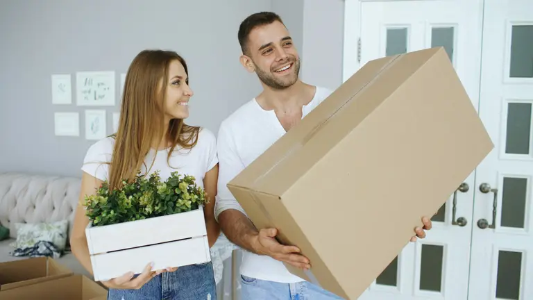 Smiling couple carrying a cardboard box and a plant into their new home
